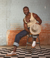 A man in a beige tank top and jeans sits on a stool, holding a hat, against a brick and painted wall with a checkered floor. He completes his relaxed look with the Freestyle sneakers by Bed|Stü, adding effortless style to the scene.