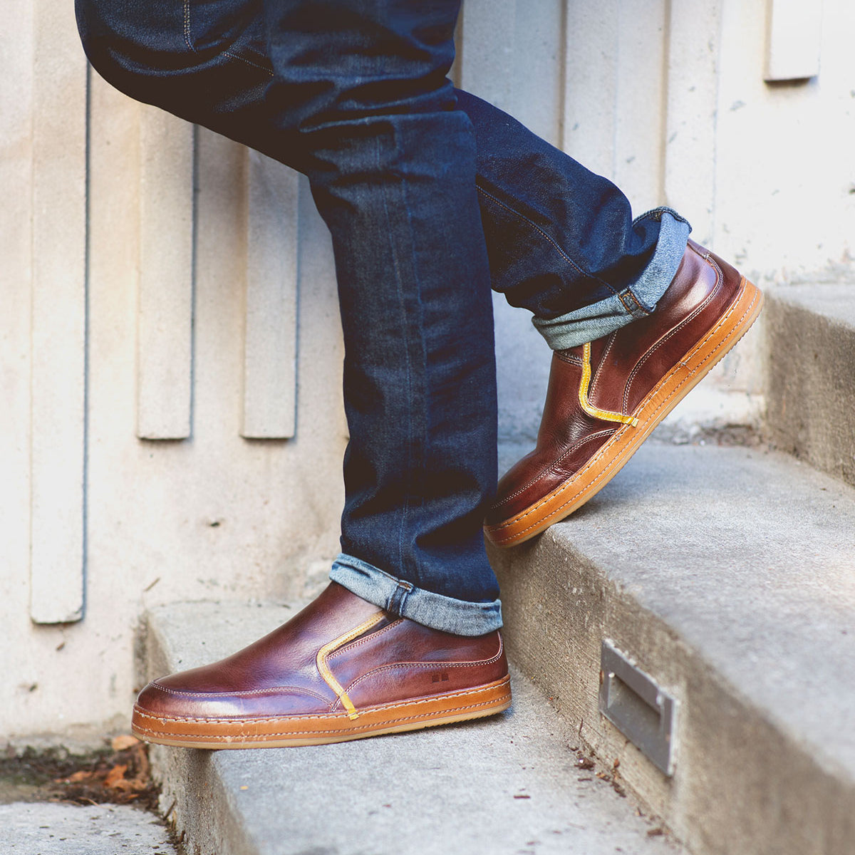 Wearing Bed|Stü Freestyle slip-on sneakers with brown leather trim and cuffed dark blue jeans, the person stands on concrete steps beside a beige wall.