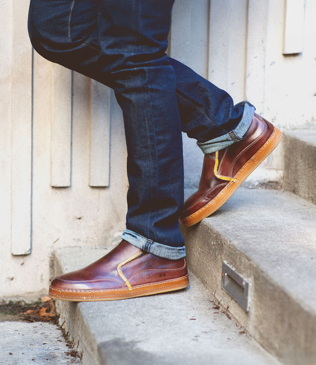 Wearing Bed|Stü Freestyle slip-on sneakers with brown leather trim and cuffed dark blue jeans, the person stands on concrete steps beside a beige wall.