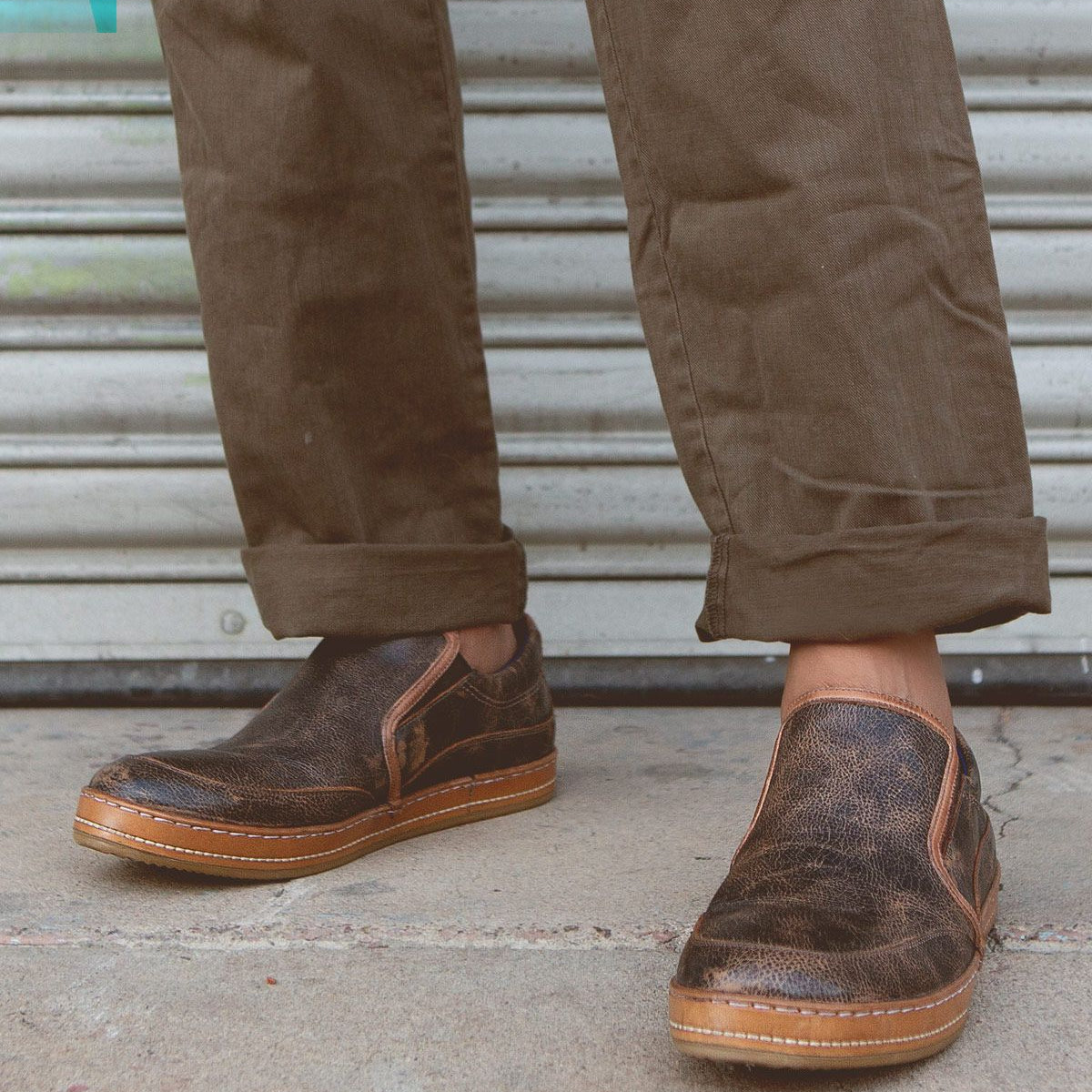A person wearing brown pants and the Bed|Stü Freestyle slip-on sneaker with leather trim stands on a concrete surface in front of a metal shutter.