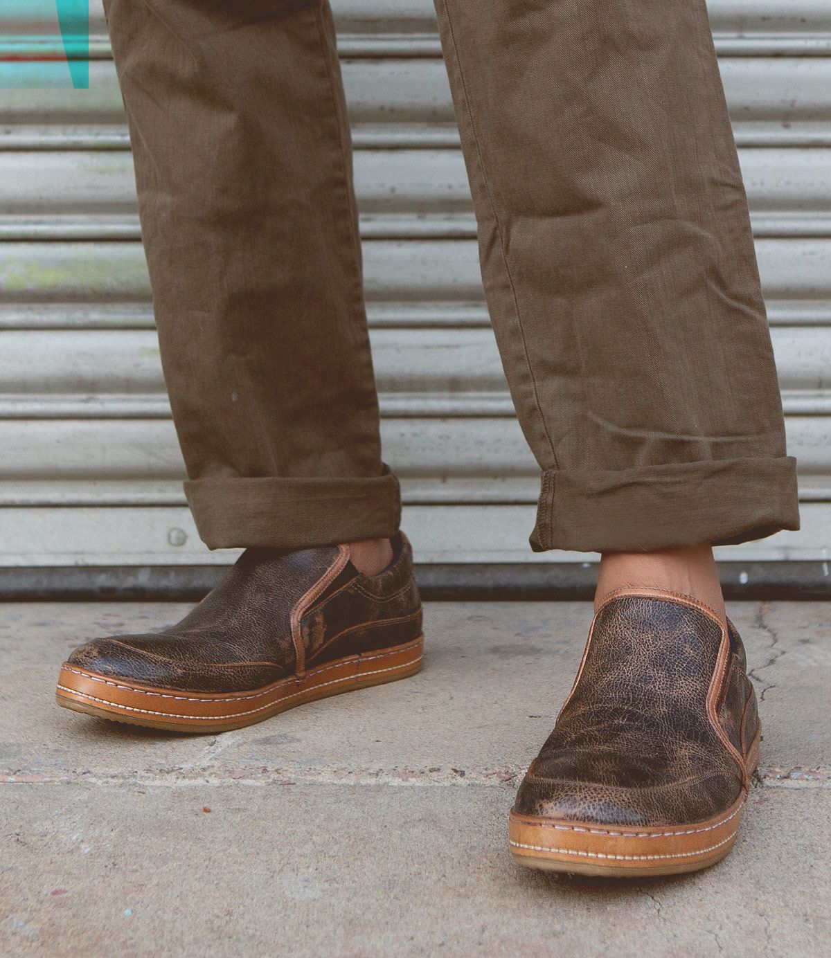 A person wearing brown pants and the Bed|Stü Freestyle slip-on sneaker with leather trim stands on a concrete surface in front of a metal shutter.