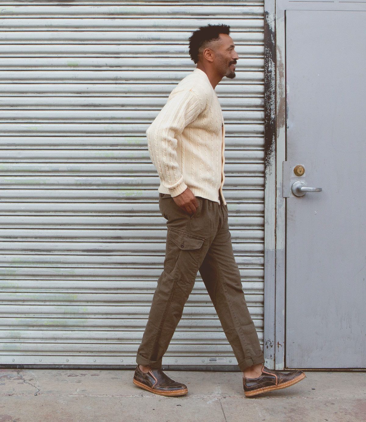 A man in a cream cardigan, olive cargo pants, and dark shoes—styled with the Bed|Stü Freestyle comfortable slip-on sneakers featuring subtle leather trim—walks past a gray metal garage door and a gray door with a round handle.