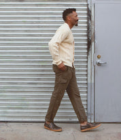 A man in a cream cardigan, olive cargo pants, and dark shoes—styled with the Bed|Stü Freestyle comfortable slip-on sneakers featuring subtle leather trim—walks past a gray metal garage door and a gray door with a round handle.