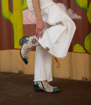 Standing in front of a colorful mural adorned with cactus and flower designs, a person in frayed white pants adjusts the Bed|Stü Frio—Western bootie ankle boots detailed with antiqued brass studs.