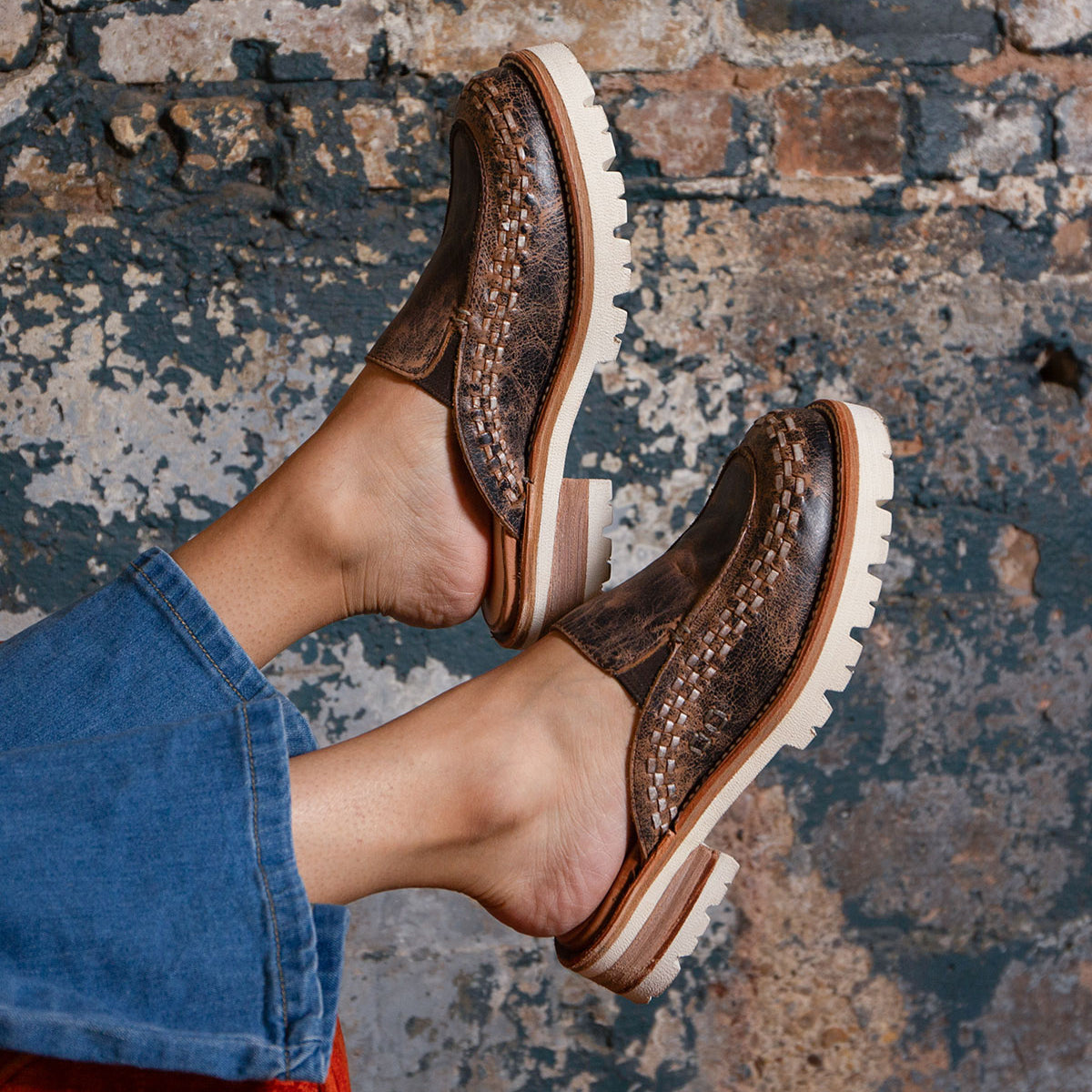 A person wearing the Fuel handwoven leather slip-on shoes by Bed|Stü, featuring XL EXTRALIGHT outsoles and light-colored soles, paired with blue denim pants, stands in front of a textured, weathered brick wall background.