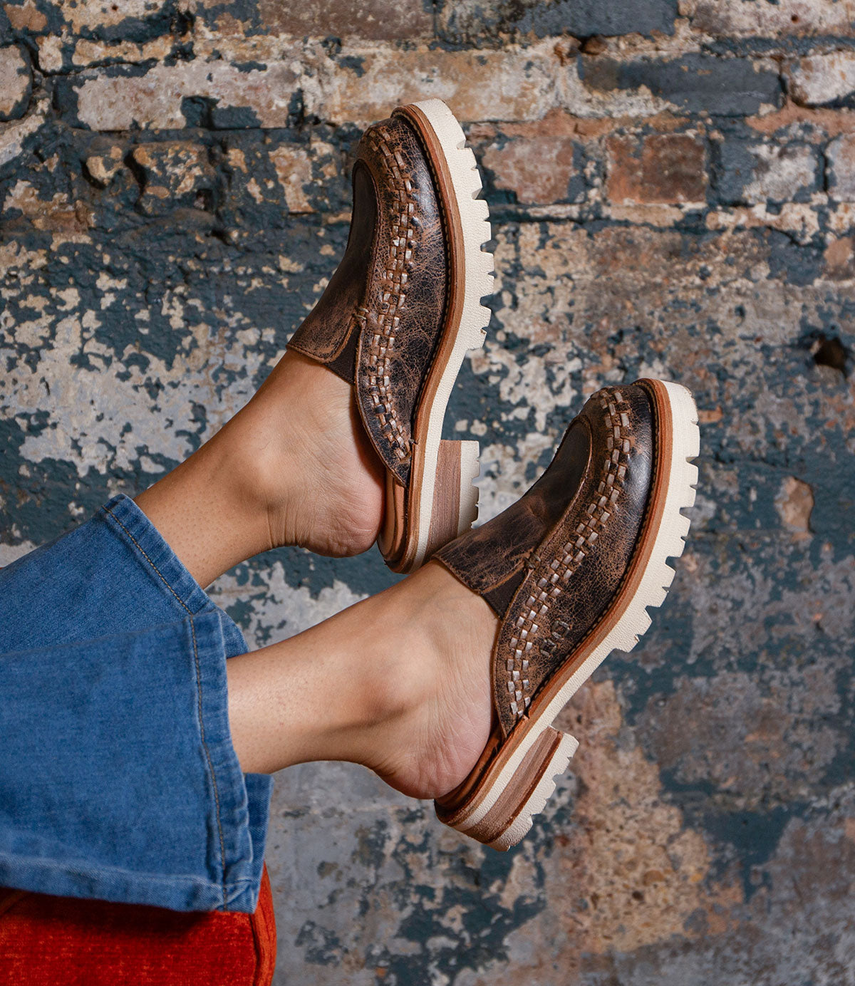 A person wearing the Fuel handwoven leather slip-on shoes by Bed|Stü, featuring XL EXTRALIGHT outsoles and light-colored soles, paired with blue denim pants, stands in front of a textured, weathered brick wall background.
