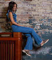 A woman dressed in a blue denim outfit sits sideways atop a vintage wooden radio against a weathered brick wall, smiling with her feet lifted off the ground, her look finished with Bed|Stü’s Fuel clogs—lightweight footwear crafted from handwoven leather.