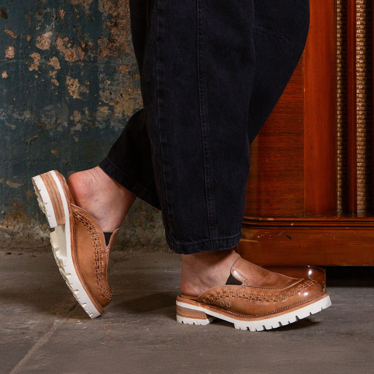 A person wearing black pants and Bed|Stü Fuel brown handwoven leather slip-on shoes with white XL EXTRALIGHT® outsoles stands indoors on a concrete floor near a wooden cabinet.
