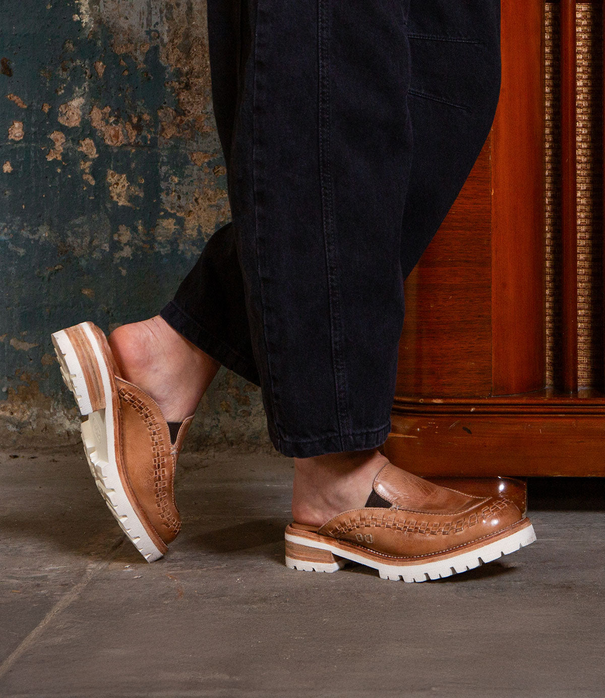 A person wearing black pants and Bed|Stü Fuel brown handwoven leather slip-on shoes with white XL EXTRALIGHT® outsoles stands indoors on a concrete floor near a wooden cabinet.