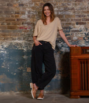 A woman stands against a worn brick wall, smiling, wearing a beige t-shirt and black pants. On her feet are Bed|Stü's Fuel sandals in brown, featuring handwoven leather details and an XL EXTRALIGHT outsole. One hand rests on a vintage wooden radio.