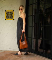 A woman in a strapless black dress, sunglasses, and sandals with a cushioned footbed stands near a glass door on a tiled patio, holding a brown bag. Her comfortable Bed|Stü Gailee leather slide sandals complete the chic and effortless look.