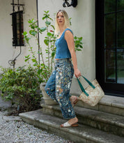 A woman wearing a sleeveless blue top, patterned jeans, and comfortable sandals with a cushioned footbed stands on outdoor steps holding the large beige Bed|Stü Gailee tote bag. Shrubs and a window can be seen in the background.