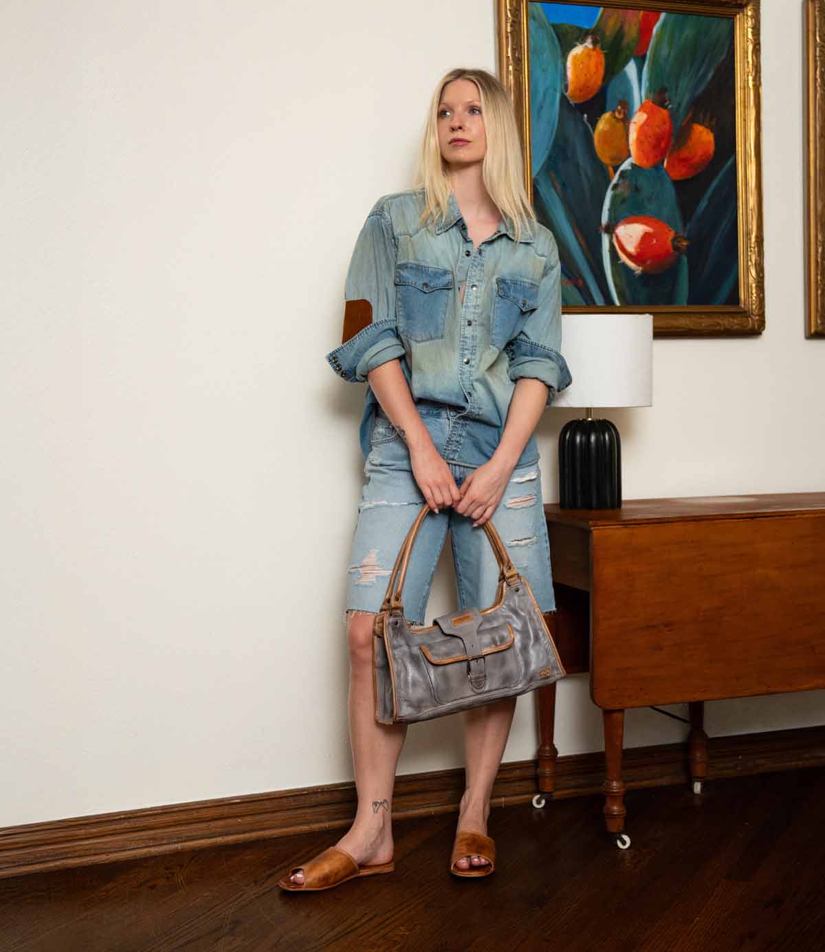 A woman stands indoors in front of a table and framed artwork, wearing a denim shirt, ripped denim shorts, and the Bed|Stü Gailee comfortable brown leather slide sandals with a cushioned footbed, while holding a gray handbag.