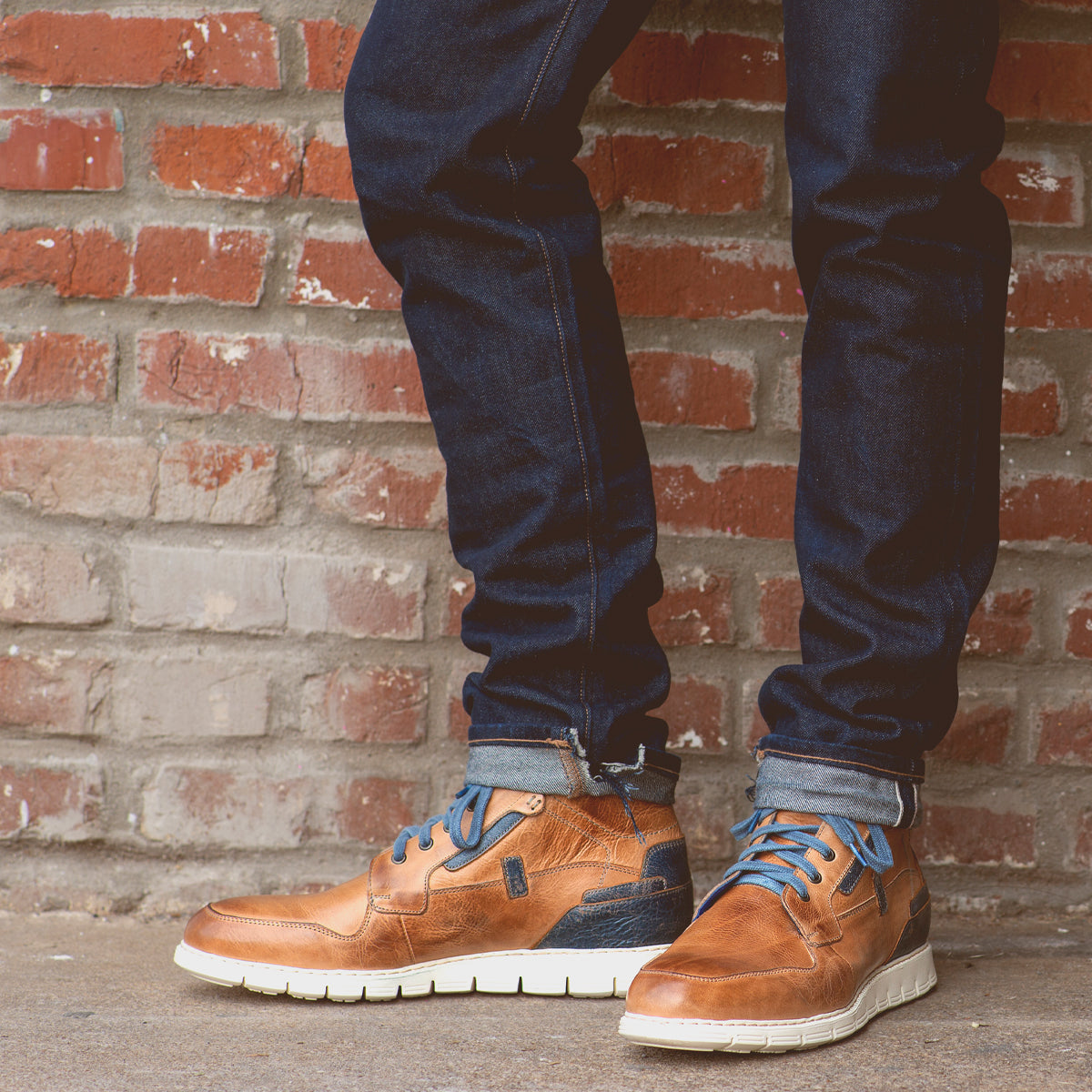 A person wearing cuffed dark jeans and Bed|Stü's Galahad brown leather sneakers—with a lightweight sole and blue laces—stands in front of a brick wall.