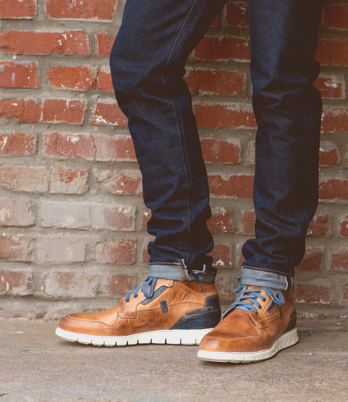 A person wearing cuffed dark jeans and Bed|Stü's Galahad brown leather sneakers—with a lightweight sole and blue laces—stands in front of a brick wall.
