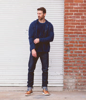 A man stands in front of a white garage door and red brick wall, wearing a navy blue jacket, dark jeans, and Bed|Stü Galahad tan high-top sneakers with a lightweight sole, looking to the side with one hand on his jacket zipper.