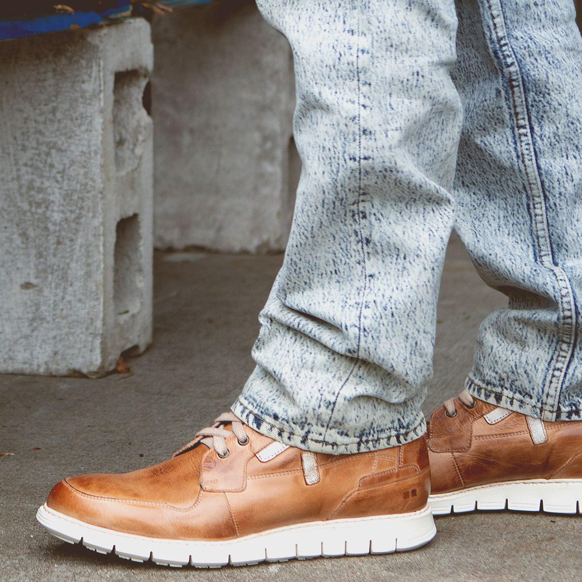 A person wearing light blue acid-wash jeans and Bed|Stü’s Galahad tan leather sneakers with a lightweight sole stands on a concrete surface near a cinder block.