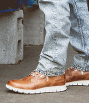 A person wearing light blue acid-wash jeans and Bed|Stü’s Galahad tan leather sneakers with a lightweight sole stands on a concrete surface near a cinder block.