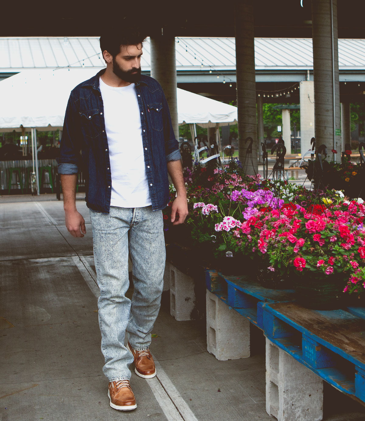A man in casual jeans and a denim shirt stands near trays of colorful flowers at an outdoor market, sporting the Bed|Stü Galahad leather sneakers with a lightweight sole for all-day comfort.