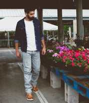 A man in casual jeans and a denim shirt stands near trays of colorful flowers at an outdoor market, sporting the Bed|Stü Galahad leather sneakers with a lightweight sole for all-day comfort.