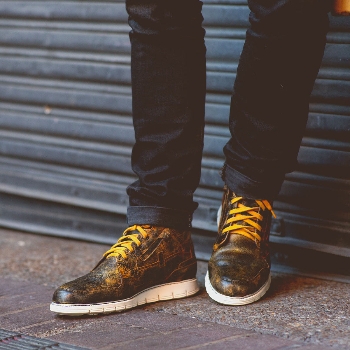 A person wearing black pants and Bed|Stü's Galahad distressed black high-top sneakers with yellow laces and a lightweight sole stands on a sidewalk in front of a metal shutter.