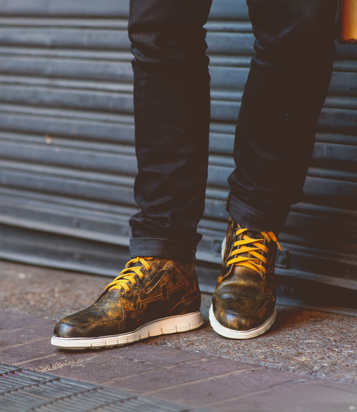 A person wearing black pants and Bed|Stü's Galahad distressed black high-top sneakers with yellow laces and a lightweight sole stands on a sidewalk in front of a metal shutter.