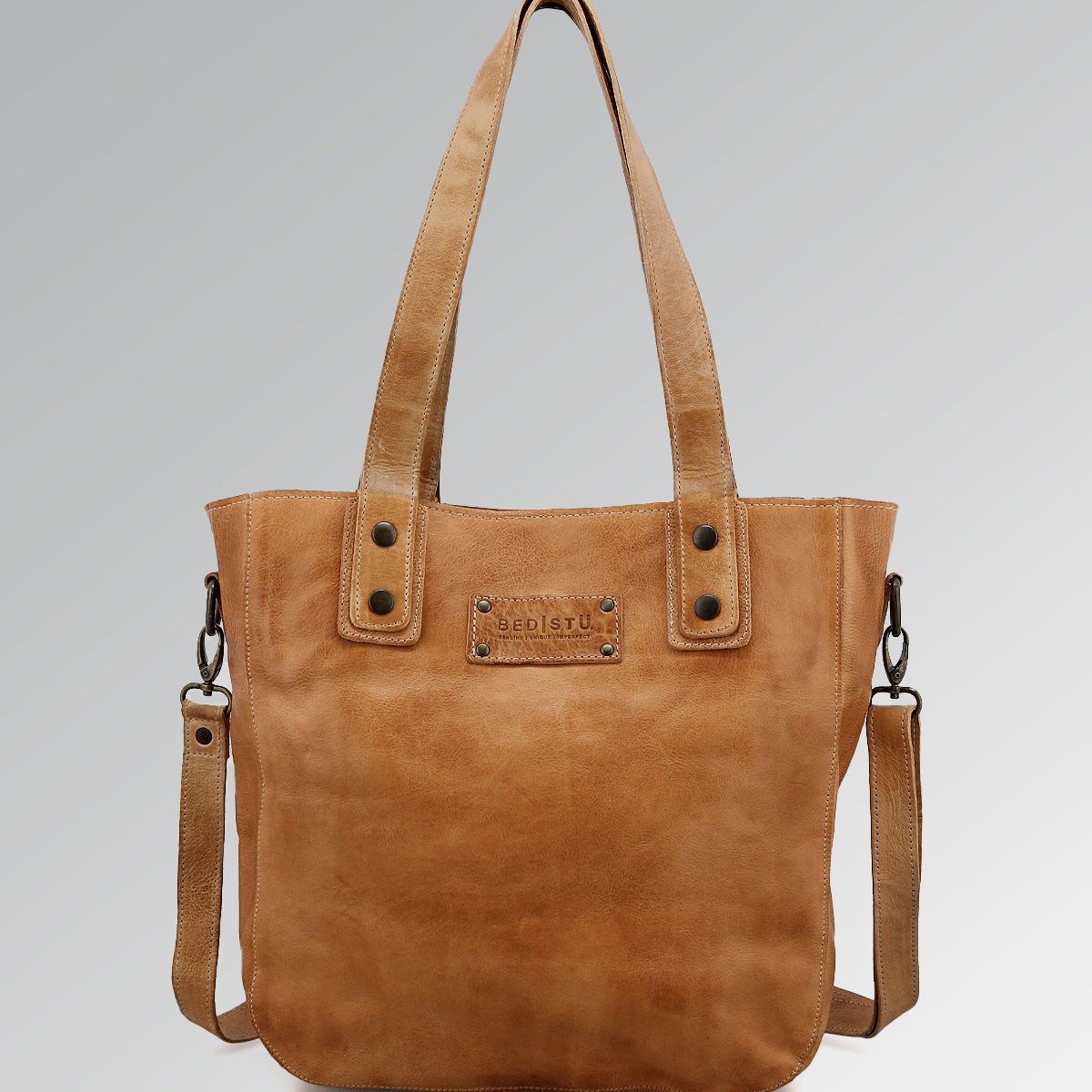 The Studio Gaviota II Body by Bed|Stü is a light brown leather tote bag featuring dual handles, a detachable shoulder strap, and a rectangular stitched patch on the front. It is displayed against a plain light gray gradient background.
