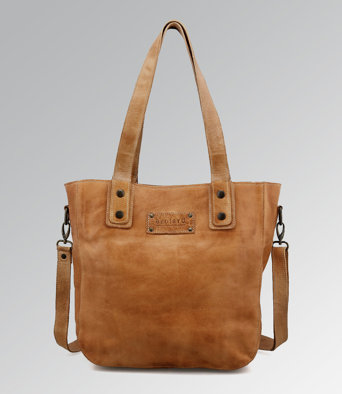 The Studio Gaviota II Body by Bed|Stü is a light brown leather tote bag featuring dual handles, a detachable shoulder strap, and a rectangular stitched patch on the front. It is displayed against a plain light gray gradient background.