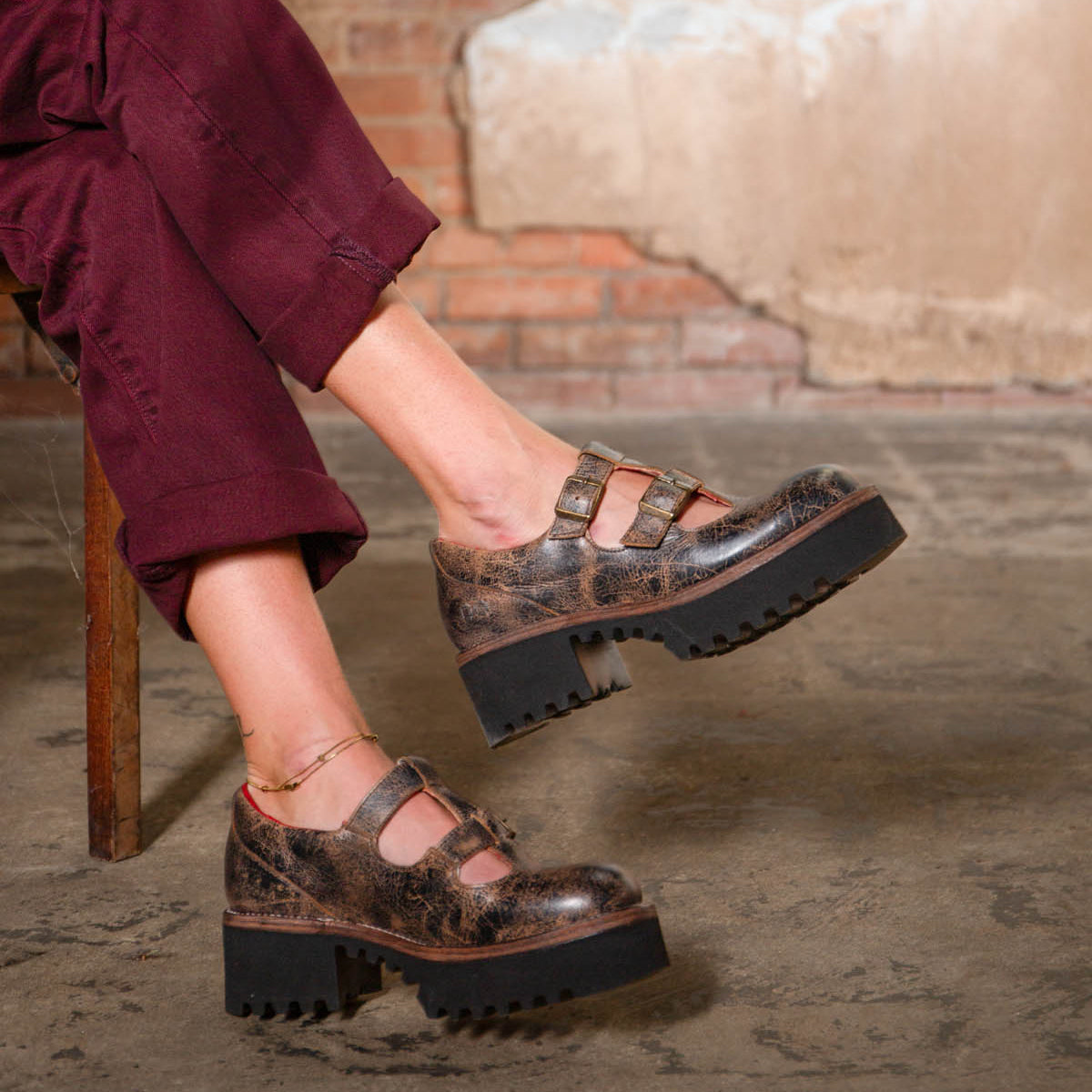 A person wearing burgundy pants and the Bed|Stü Gear chunky brown women’s shoe, featuring a double-strap design and XL Extralight sole, sits cross-legged on a wooden chair against a textured wall and concrete floor.