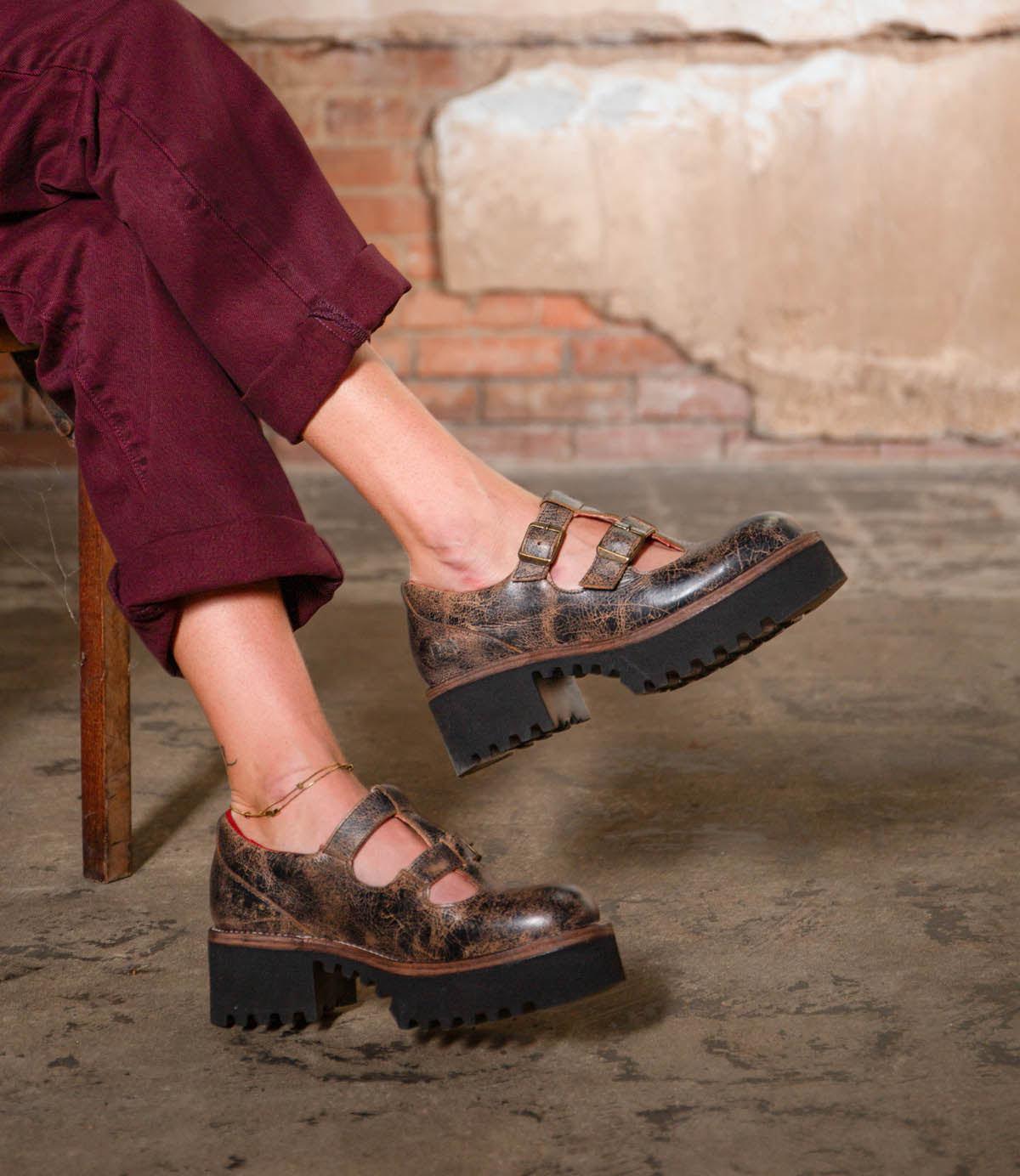 A person wearing burgundy pants and the Bed|Stü Gear chunky brown women’s shoe, featuring a double-strap design and XL Extralight sole, sits cross-legged on a wooden chair against a textured wall and concrete floor.