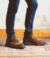 A person wearing the Bed|Stü Goer dark brown oiled suede leather cap-toe boots and blue jeans stands on a patterned rug, with a wooden baseboard and light-colored wall in the background.