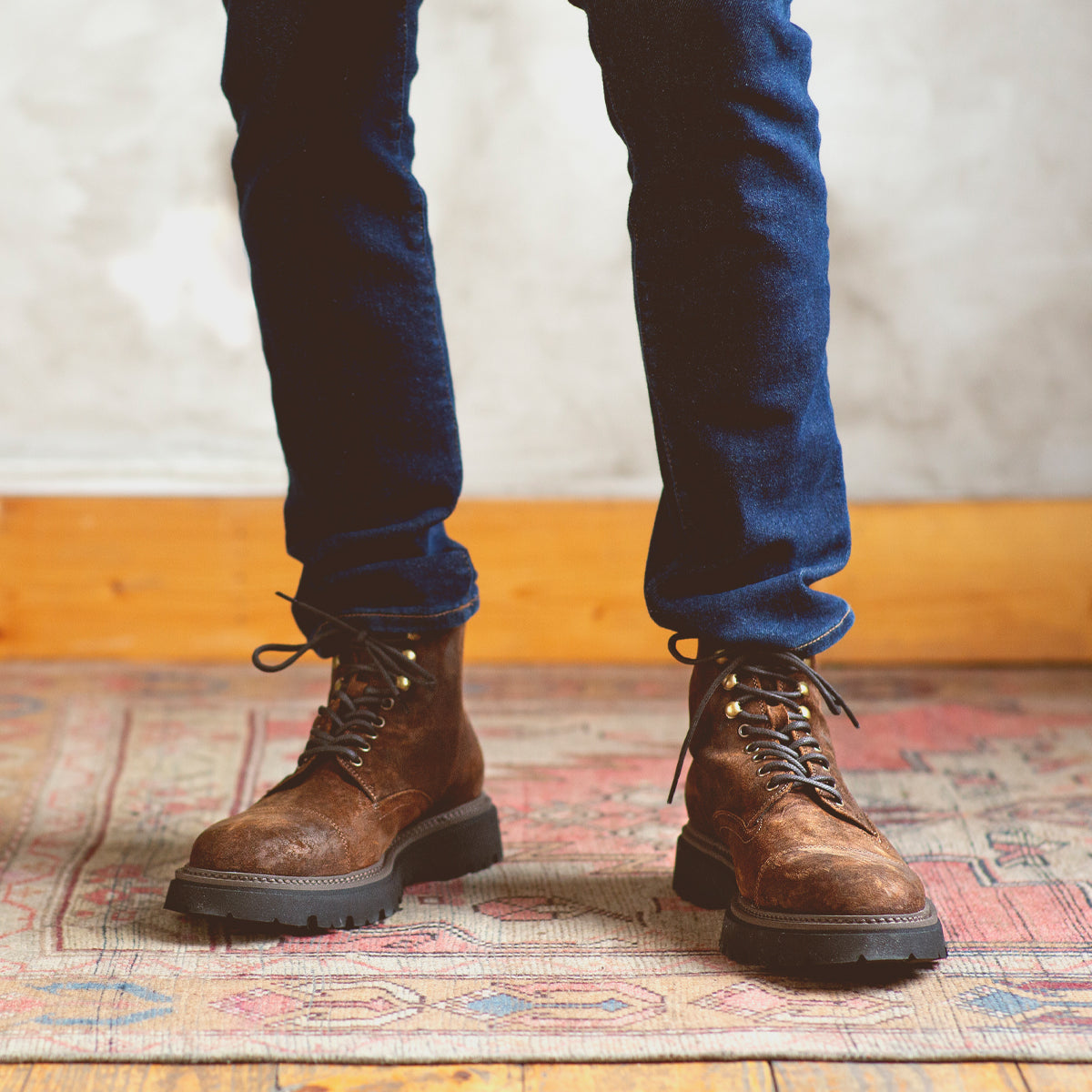 A person wearing dark blue jeans and Bed|Stü Goer brown Italian work-style cap-toe boots stands on a patterned rug with a wooden floor and light-colored wall in the background.