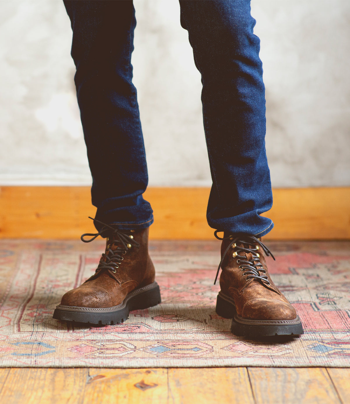 A person wearing dark blue jeans and Bed|Stü Goer brown Italian work-style cap-toe boots stands on a patterned rug with a wooden floor and light-colored wall in the background.