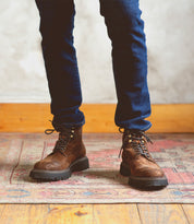 A person wearing dark blue jeans and Bed|Stü Goer brown Italian work-style cap-toe boots stands on a patterned rug with a wooden floor and light-colored wall in the background.