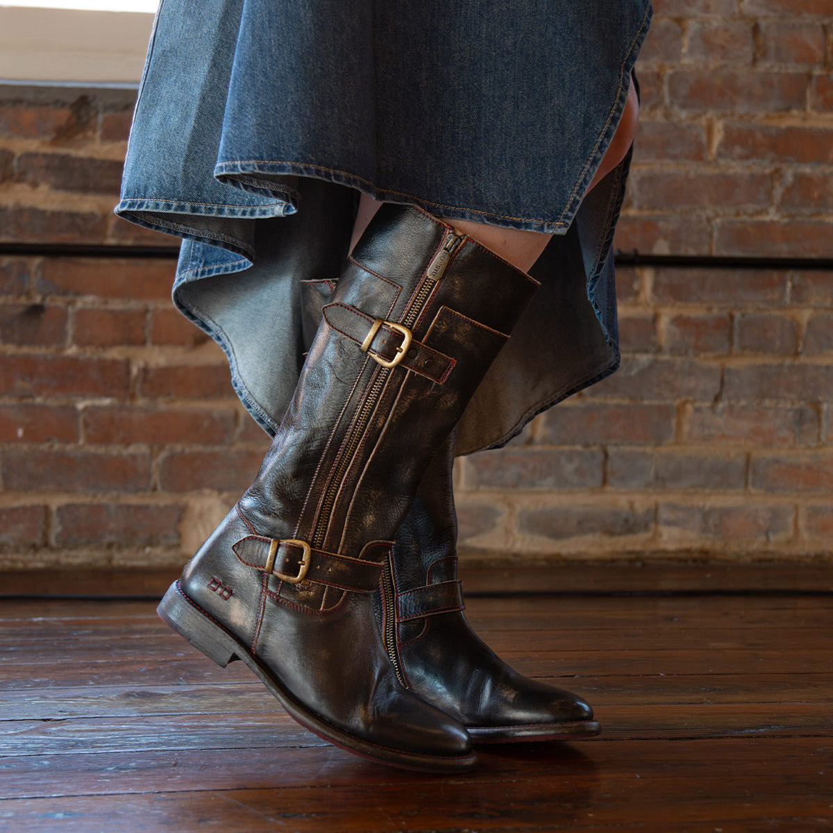 A person wearing a high-low hem denim skirt and the Bed|Stü Gogo Lug dark brown knee-high leather boots with buckles and a sturdy lug outsole, standing on a wooden floor in front of a brick wall.