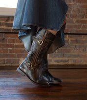 A person wearing a high-low hem denim skirt and the Bed|Stü Gogo Lug dark brown knee-high leather boots with buckles and a sturdy lug outsole, standing on a wooden floor in front of a brick wall.
