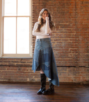 A person stands indoors near a window, wearing a white long-sleeve shirt, an asymmetrical denim skirt, and black Bed|Stü Gogo Lug mid-calf leather boots with a lug outsole, one hand touching their face.