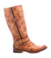 The Bed|Stü Gogo Lug is a brown leather knee-high engineer boot featuring a side zipper, two decorative buckles, and a low wooden heel, complete with a durable lug outsole; pictured in profile against a white background.