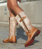 A person wearing Bed|Stü Gogo Lug knee-high tan leather boots with a lug outsole, zippers, and buckles stands on a concrete surface.