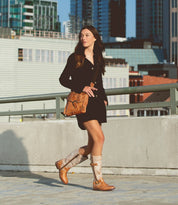 A woman wearing a black dress, tan shoulder bag, and Bed|Stü Gogo Lug mid-calf leather boots with a rugged lug outsole walks on a rooftop parking structure with city buildings in the background.
