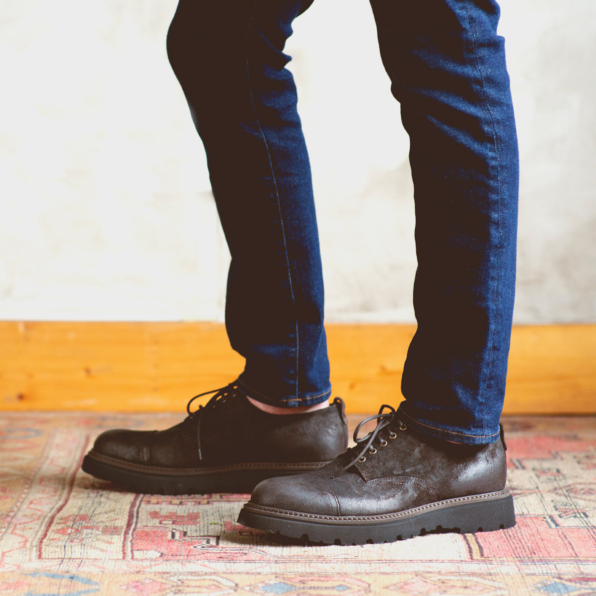 A person wearing dark blue jeans and Bed|Stü Grant black Italian leather shoes stands on a patterned rug, with a light-colored wall and wooden trim in the background.