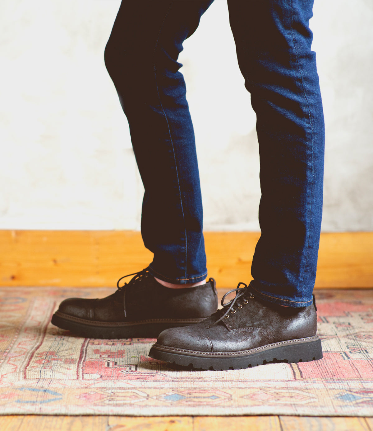 A person wearing dark blue jeans and Bed|Stü Grant black Italian leather shoes stands on a patterned rug, with a light-colored wall and wooden trim in the background.