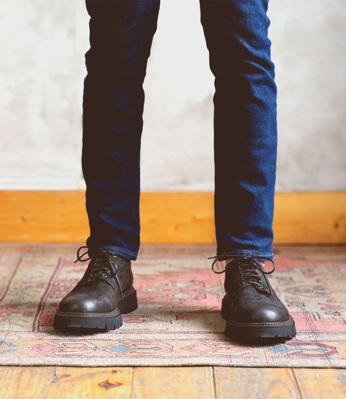 A person wearing the Grant black lace-up cap-toe derby boots by Bed|Stü and blue jeans stands on a patterned rug, with a wooden baseboard and white wall in the background.