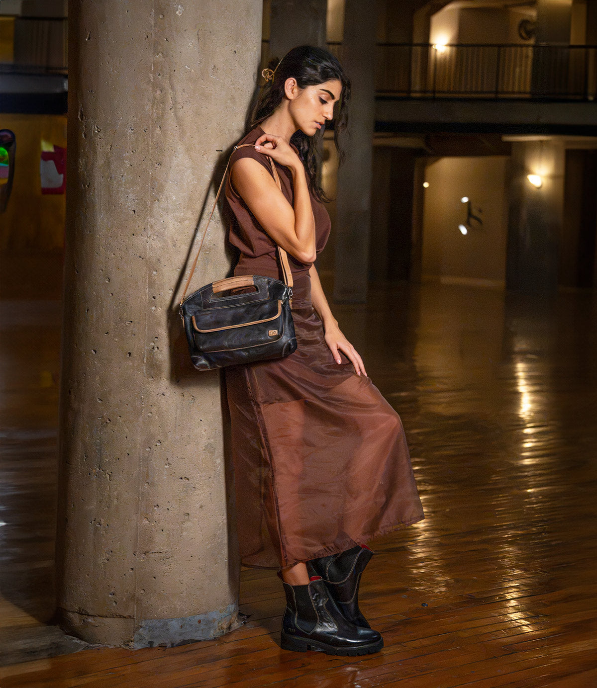 A woman in a brown dress and black boots leans against a concrete column, carrying a black and tan full-grain leather Greenway crossbody bag with a zip-top closure by Bed|Stü.