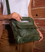A person stands in front of a brick wall, wearing a white top and brown pants, holding the Bed|Stü Greenway—a green full-grain leather handbag featuring an adjustable crossbody strap and zip-top closure.