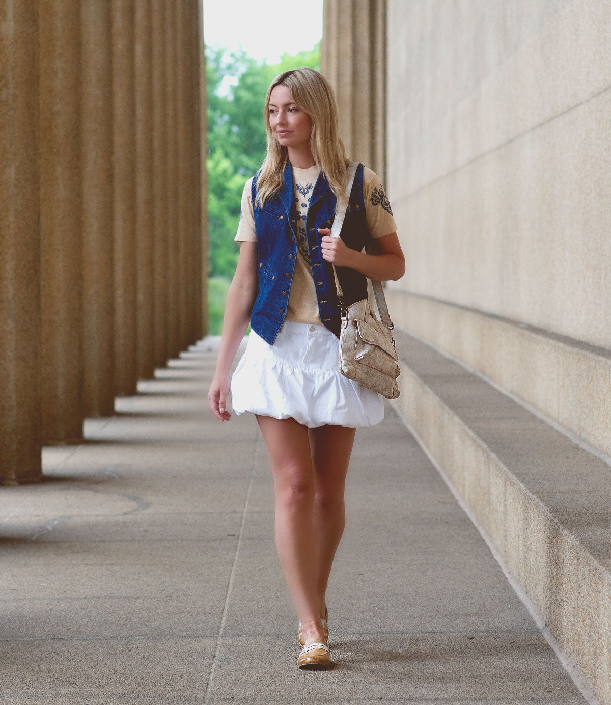 A woman in a denim vest, cream shirt, and white skirt walks along a colonnade with the Bed|Stü Greenway—a chic full-grain leather shoulder bag featuring a crossbody strap—looking ahead.