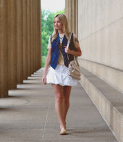 A woman in a denim vest, cream shirt, and white skirt walks along a colonnade with the Bed|Stü Greenway—a chic full-grain leather shoulder bag featuring a crossbody strap—looking ahead.