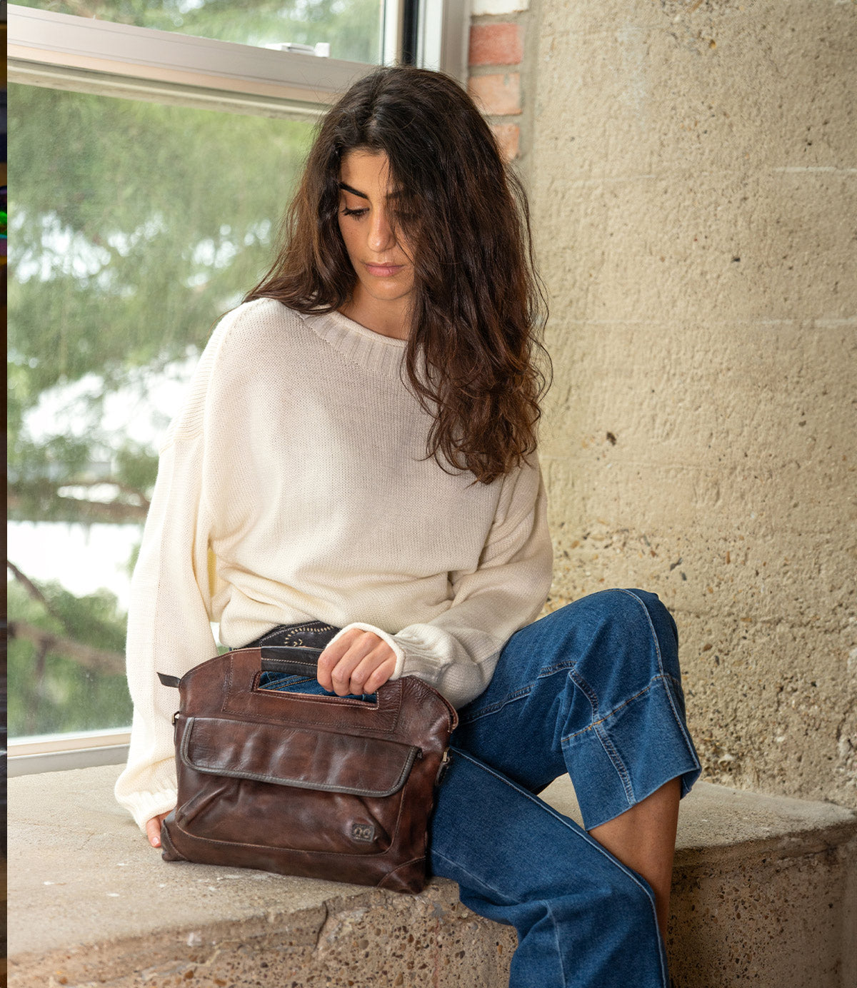 A woman in a cream sweater and blue jeans sits by a window, holding a Bed|Stü Greenway full-grain leather handbag with a zip-top closure.