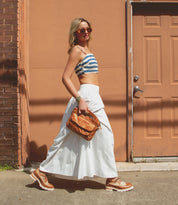 A woman in a striped crop top, white skirt, and tan shoes walks on a sidewalk carrying the Bed|Stü Greenway brown bag with a zip-top closure, passing an orange door.