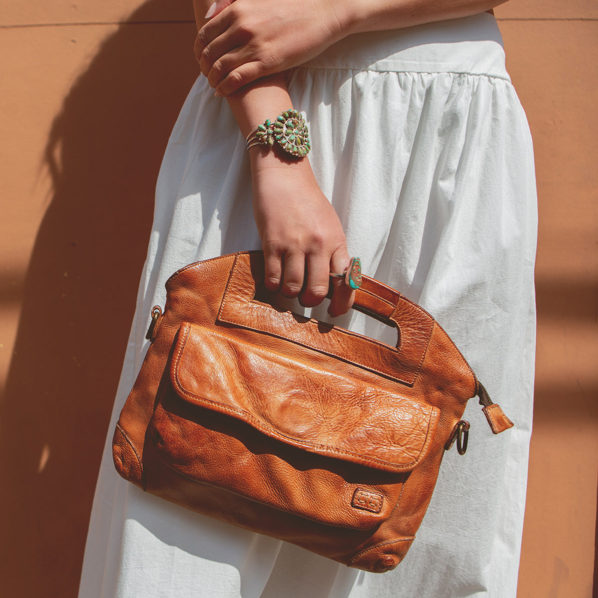 A person in a white skirt holds the Bed|Stü Greenway brown full-grain leather handbag with cutout handles and a zip-top closure, accessorizing with a large green ring and matching bracelet while standing against a peach-colored background.